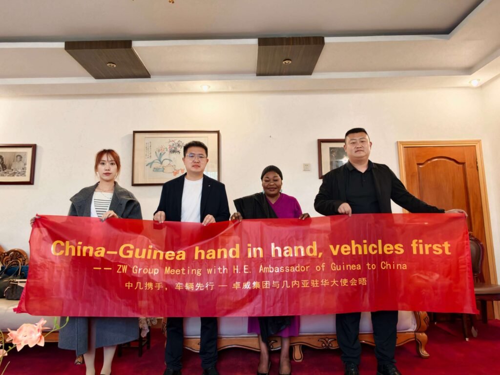 Chairman Ivan Sun and General Manager Pierce Guo of ZW Group stand with H.E. Aminata Koita, Ambassador of Guinea to China, at the Embassy, holding a banner "China-Guinea hand in hand, vehicles first."